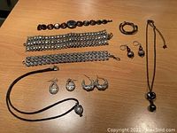Photo shows a collection of costume jewelry laid out on a wooden surface including two wide silver-tone chain-link bracelets, various pairs of earrings and a black cord necklace with a silver-tone pendant.