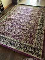 Photo showing large rectangular floor mat with burgundy base and beige floral design bordered by a black and beige floral patterned edge.