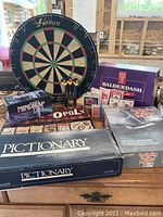 Photo showing Wiflyte dartboard with six darts and multiple board games including Pictionary (First Edition), Mindtrap, Balderdash, and a mystery game, plus packs of playing cards.