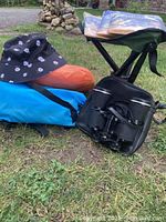 Outdoor setting photo showing two inflatable lounge chairs (orange and blue), a small blue and green Coleman camp seat, black binoculars in a case, and a black reversible sun hat with white floral print.
