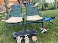 Two green metal lawn chairs with cream cushions, two hats placed on chair backs, blue bird garden wind spinner in background, and garden figurines including owl, angels, and planter in front.