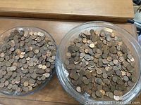 Top-down view of two clear glass bowls filled with mixed pennies sitting on a wooden table.