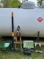 Photo showing Scotts handheld seed and fertilizer spreader, long-handled weed puller, lopper, and 8 lb sledgehammer head in front of a white propane tank