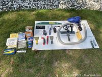 Wide shot of all items laid out on a white board on grass including drywall tools, vacuum, hose, light bulbs, and other electrical items.