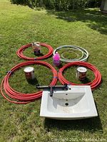 Overall lot showing four coils of PEX piping (3 red, 1 white), two cans of wood stain, jug of RV antifreeze, porcelain sink, and stove pipe ring on grass.