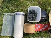 Photo showing part rolls of green and white rolls of sealant and poly sheeting, a roll of brown Resisto tape and container of green steel roofing screws on grass.