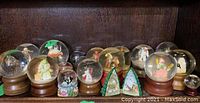 Photo of 18 snow globes arranged on a wooden shelf. Includes multiple snowmen, Christmas themed globes, and other figurines. Various sizes and bases visible.