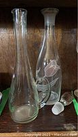 Photo showing both glass items: a clear glass decanter with handle and lid, and a tall, slender bottle with pink roses painted on it.