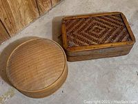 Two wooden containers on cement floor against wood panel wall, round light woven container and rectangular dark woven container.