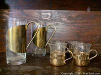 Photo showing two large and four small glass cups with brass handles on a wooden surface.