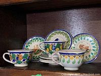 Photo of 3 cups and 6 saucers showing hand-painted colorful floral and bird patterns. Cups are positioned in front with saucers upright behind them on a shelf.