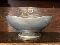 Side view of the Wilton metal bowl on a wooden surface showing its oval shape and pedestal base with worn and scratched finish.