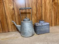 Galvanized watering can and square metal bin with lid side by side on wooden surface against wood paneling walls.