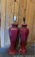 Pair of maroon ceramic table lamps shown front facing against a wooden wall background. One lamp has a white socket, the other a black socket.