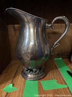 Front view of tarnished Apollo pitcher showing bulbous body, ornate flared spout edge, and sculptural base details.