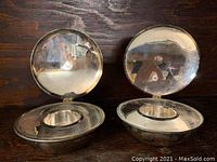 Two round, silver-tone votive candle holders shaped like shells with hinged lids, reflective surface showing some tarnishing and surface wear