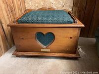Small wooden chest with hinged lid closed, featuring blue cushioned top and heart-shaped cutout on front panel with brass plate underneath.