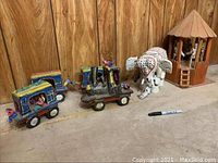 Photo of all folk art items including the elephant, train set, skeletons with hut, placed on floor against wood paneling