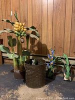 Image showing large wooden banana plant with yellow and green fruit and large green leaves, along with a metal bin and two smaller plant sculptures on a wood panel wall background.