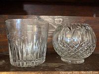 Two lead crystal vases placed side by side on a wooden surface, one cylindrical with leaf and vertical cuts, one spherical with diamond cuts.