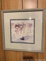 Full view of the framed wolf art print showing five wolves in snow inside a silver frame on wood panel background.