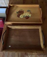 Top-down photo showing two wooden rectangular trays; the top tray is lighter wood with colorful painted fruit design and wooden side handles, the bottom tray is darker wood with black metal handles and a price tag sticker.