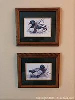 Two framed duck prints hung vertically on a wall, showing wood frames and green mats.