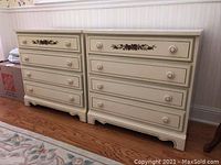 Photo of two matching cream-colored dressers with floral decorations on top drawer, showing full front view, placed side by side on hardwood floor.