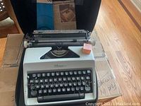 Full view of the Olympia typewriter in open carrying case with manual placed behind it and an orange box on right side.