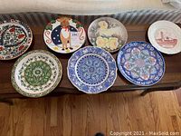 Seven decorative plates on a wooden surface, showing a variety of floral, geometric, and figure designs in multiple colors, some handmade in Greece.