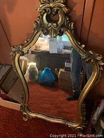 Large ornate carved gold-tone framed wall mirror leaning against a wall showing detailed carvings at the top and bottom of the frame.
