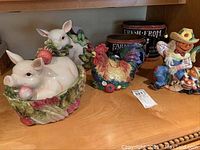 Four Fitz and Floyd ceramic pieces: a white pig nestled in vegetables, a standing pig with vegetable decorations, a colorful rooster, and a smiling farmer figurine, accompanied by two brown tins with farm lettering.