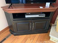 Corner black wooden entertainment center positioned in corner with open shelf holding a device and closed storage cabinet below with ornamental metal handles.