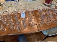 Photo showing multiple rows of clear glass stemware with gold rims on a wooden table, including wine glasses, cocktail glasses, and coupe glasses largely arranged in groups.