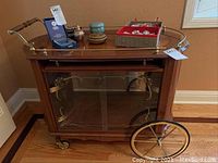 Angle view of entire tea cart showing medium brown wood frame, brass railing, large brass spoked wheels and various items on top shelf