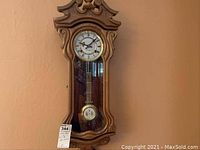 Photo of ornate wooden pendulum wall clock with pendulum visible behind glass and Roman numeral clock face.