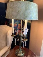 Full view of one brass and crystal table lamp with beige textured fabric shade, placed next to a chair.