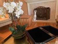 White faux orchid plant arrangement in brown ceramic pot on gold surface near wooden box and fireplace.
