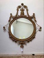 Full view of large round decorative wall mirror hanging on a white wall, showing the ornate gold toned frame with floral and beaded motifs.
