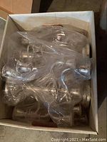 Six silver plated candelabras packed inside a cardboard box partially covered with plastic wrap.