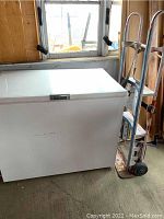 White chest freezer with appliance dolly next to it in a wooden room.