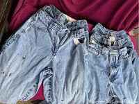 Front view of five pairs of light blue denim ladies jeans folded on a pink fabric background showing stains and wear