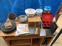 Four pairs of bookends, a wooden tea box labeled 'Bentley's Finest Teas' with lid, stack of circular coasters, metal strainer, small round makeup mirror, three plastic dehydrator trays, two Disney plastic buckets, and a congratulations banner on a cart against blue backdrop