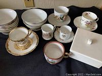 Antique demitasse cups, saucers, small bowls and other assorted china items displayed on a fabric surface.