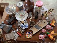 Overview of lot contents on a wooden table showing antique kitchen tools, ceramic jugs, metal grater, porcelain clogs, decorative fish and ducks, glass bottles, and a candle holder.