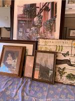 Seven framed art pieces arranged leaning against a wall and furniture, showing a variety of frames and artwork including floral, nature, and portrait subjects.