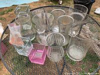 Image of 11 assorted glass vases on a metal mesh table outdoors. Includes various shapes, clear and pink glass.