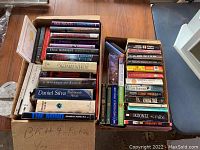 View of two boxes filled with books on a wooden table, showing mostly hardcover books and paperbacks, with a pair of concrete bookends nearby.