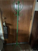 Full view of the green metal coat rack standing against a wooden door, showing its height and multiple hooks.