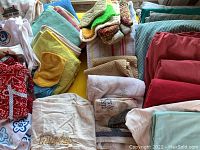 Photo showing various folded dish towels, rags, and aprons in multiple colors including red, white, yellow, and green, laid out on a surface.